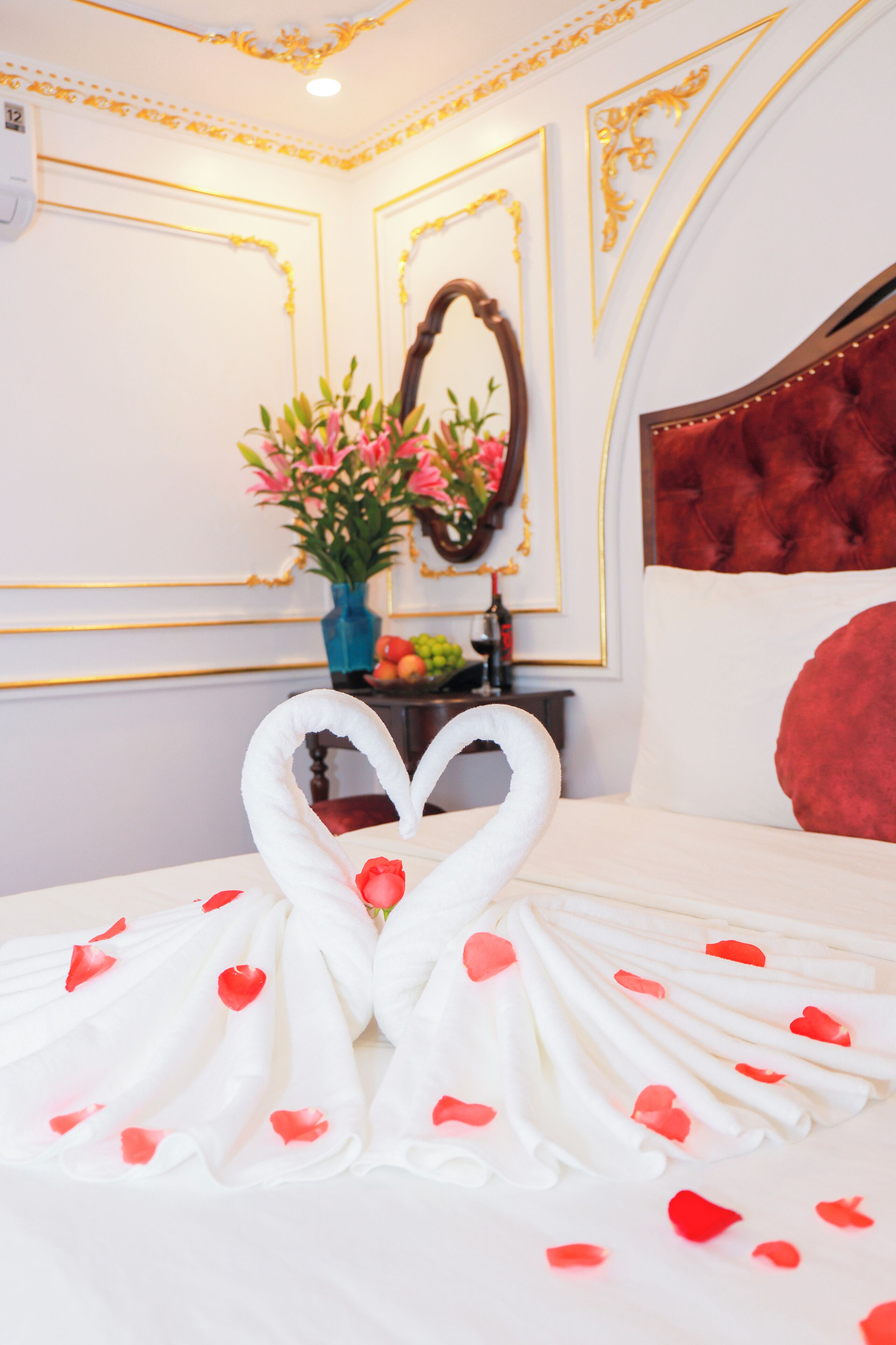 A hotel room with a bed, a mirror, a vase of flowers, and a heart-shaped towel arrangement.