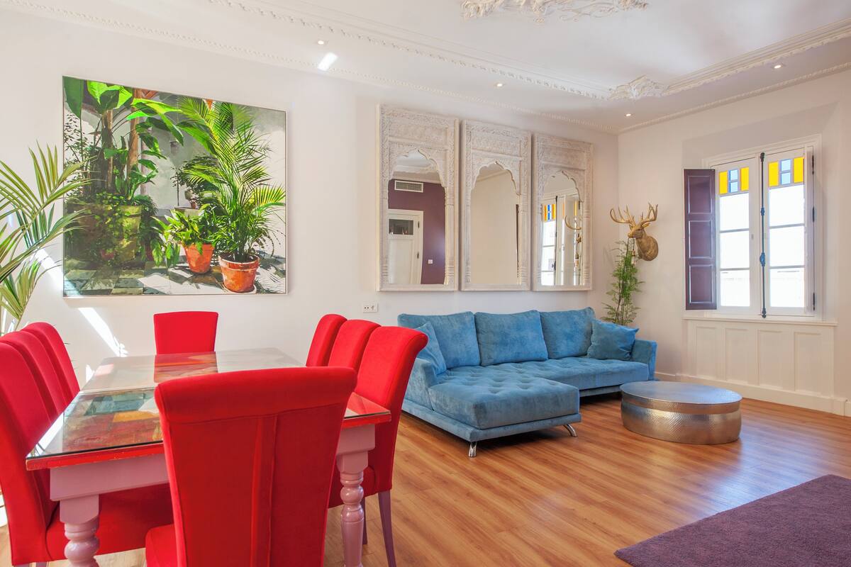 Un salon avec un canapé bleu, des fauteuils rouges, une table basse en verre et un grand tableau représentant des plantes.