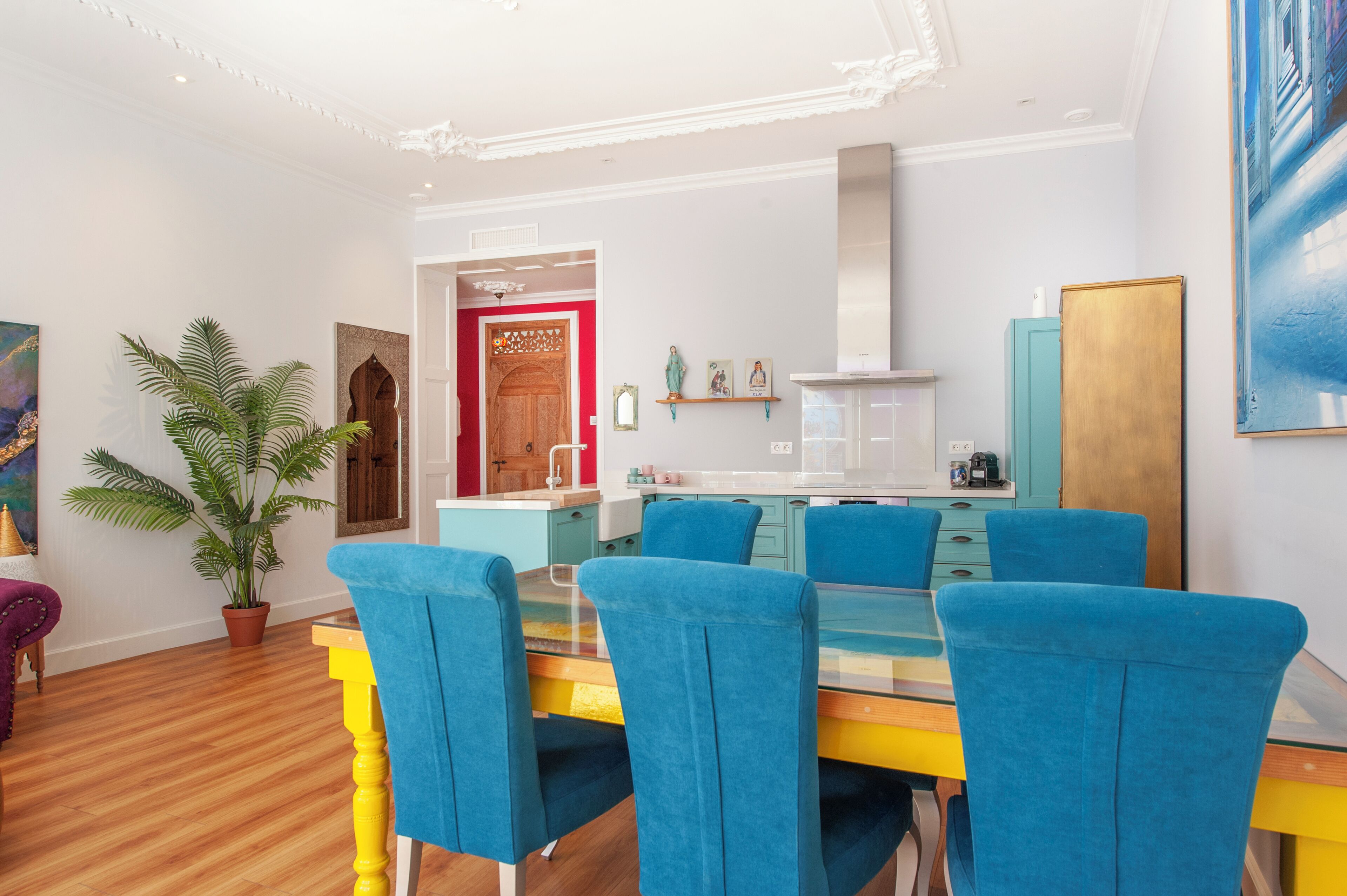 A modern dining area with a glass table and blue chairs, a potted plant, and a wooden door leading to the kitchen.
