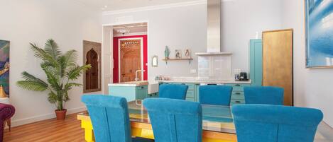A modern dining area with a glass table and blue chairs, a potted plant, and a wooden door leading to the kitchen.