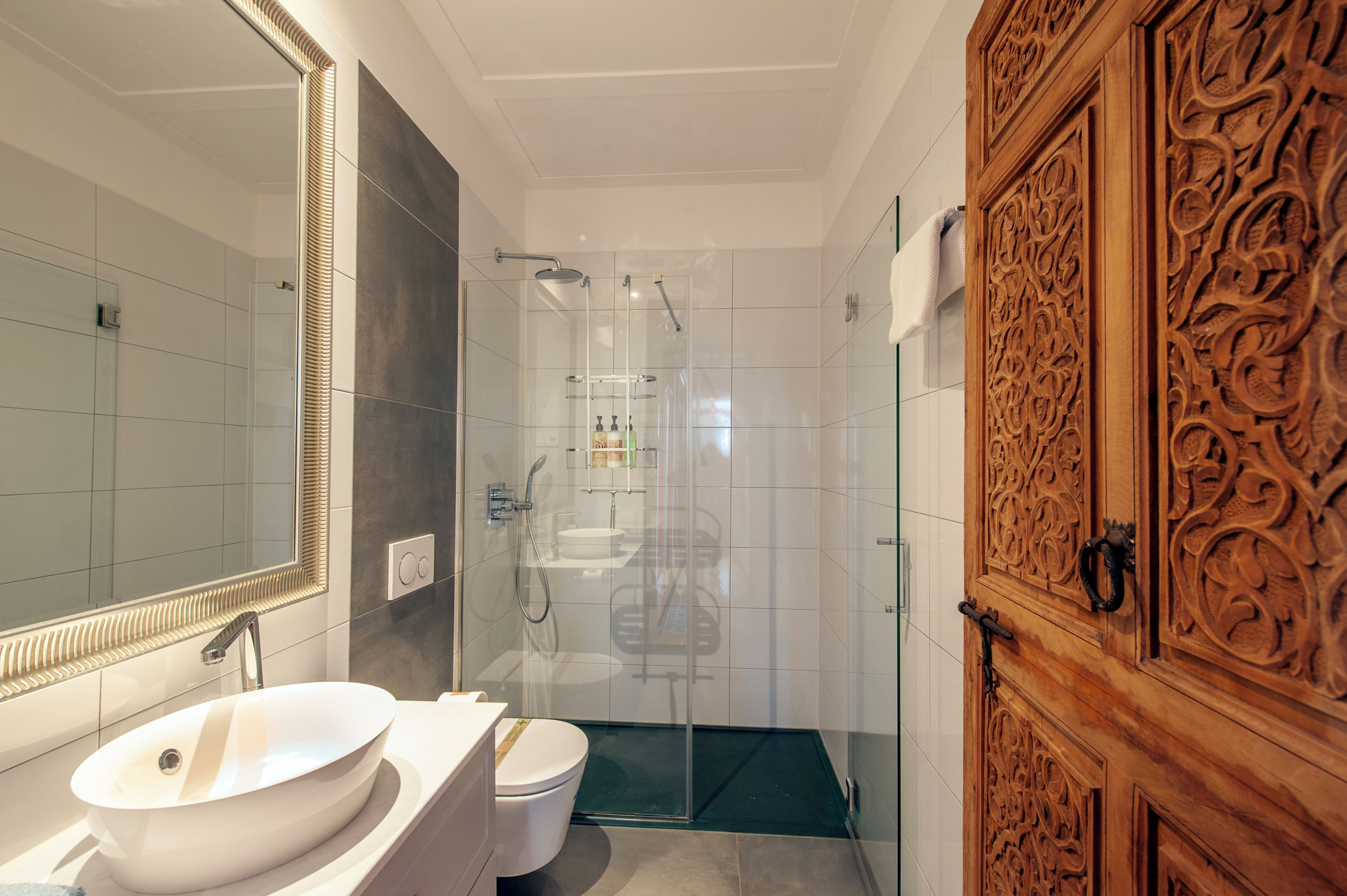 A modern bathroom with a large mirror, a white sink, a glass shower enclosure, and a wooden door with intricate carvings.