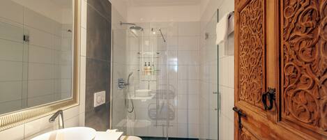 A modern bathroom with a large mirror, a white sink, a glass shower enclosure, and a wooden door with intricate carvings.