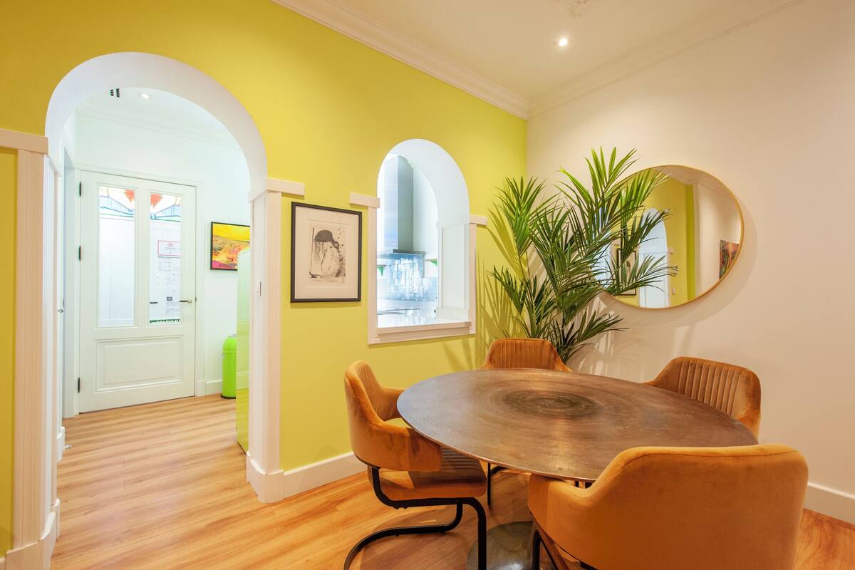 Un coin repas avec une table ronde et quatre chaises, une grande plante en pot et un miroir au mur.