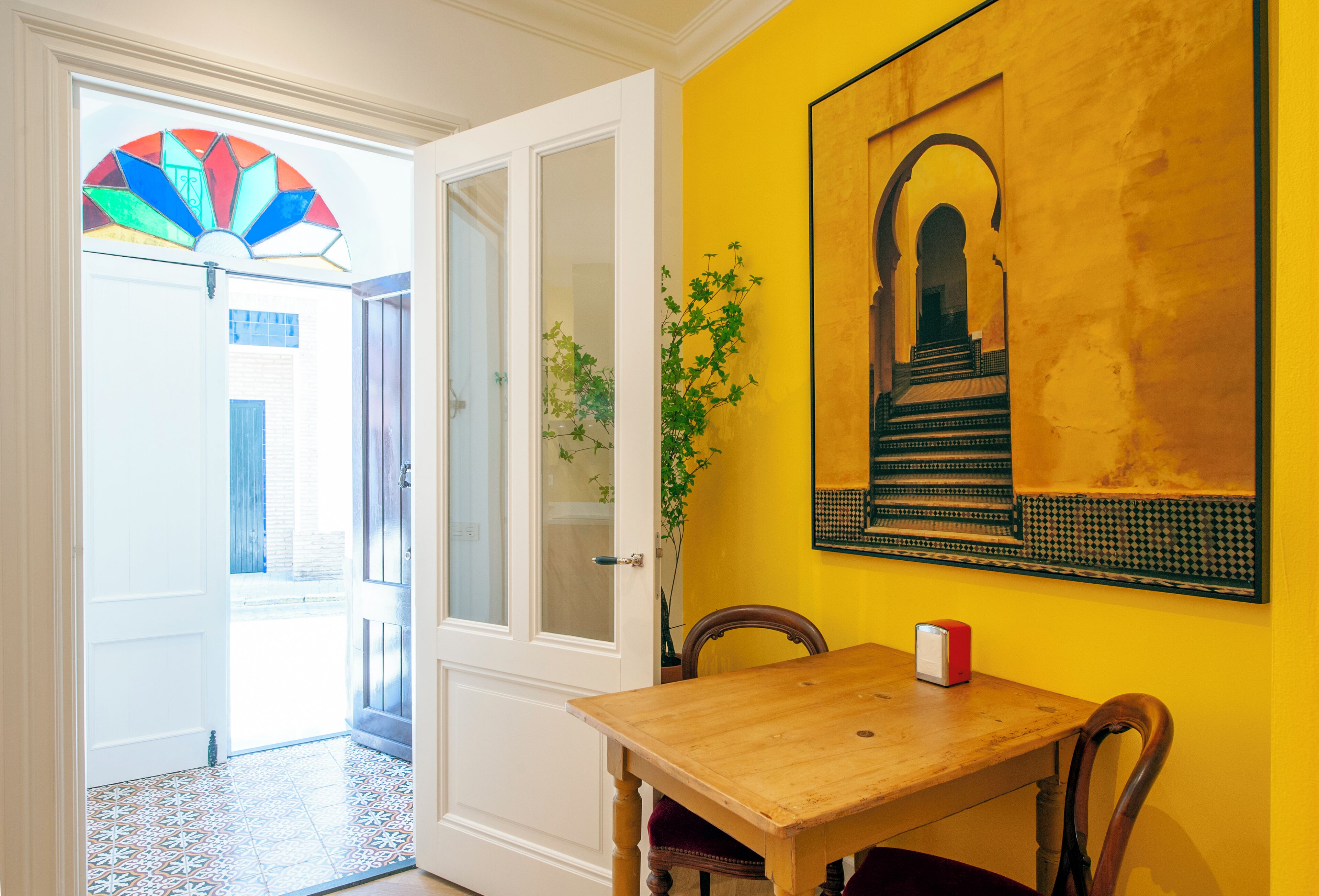 A room with a wooden table, a chair, a vase with a plant, a door with frosted glass, and a colorful stained glass window.