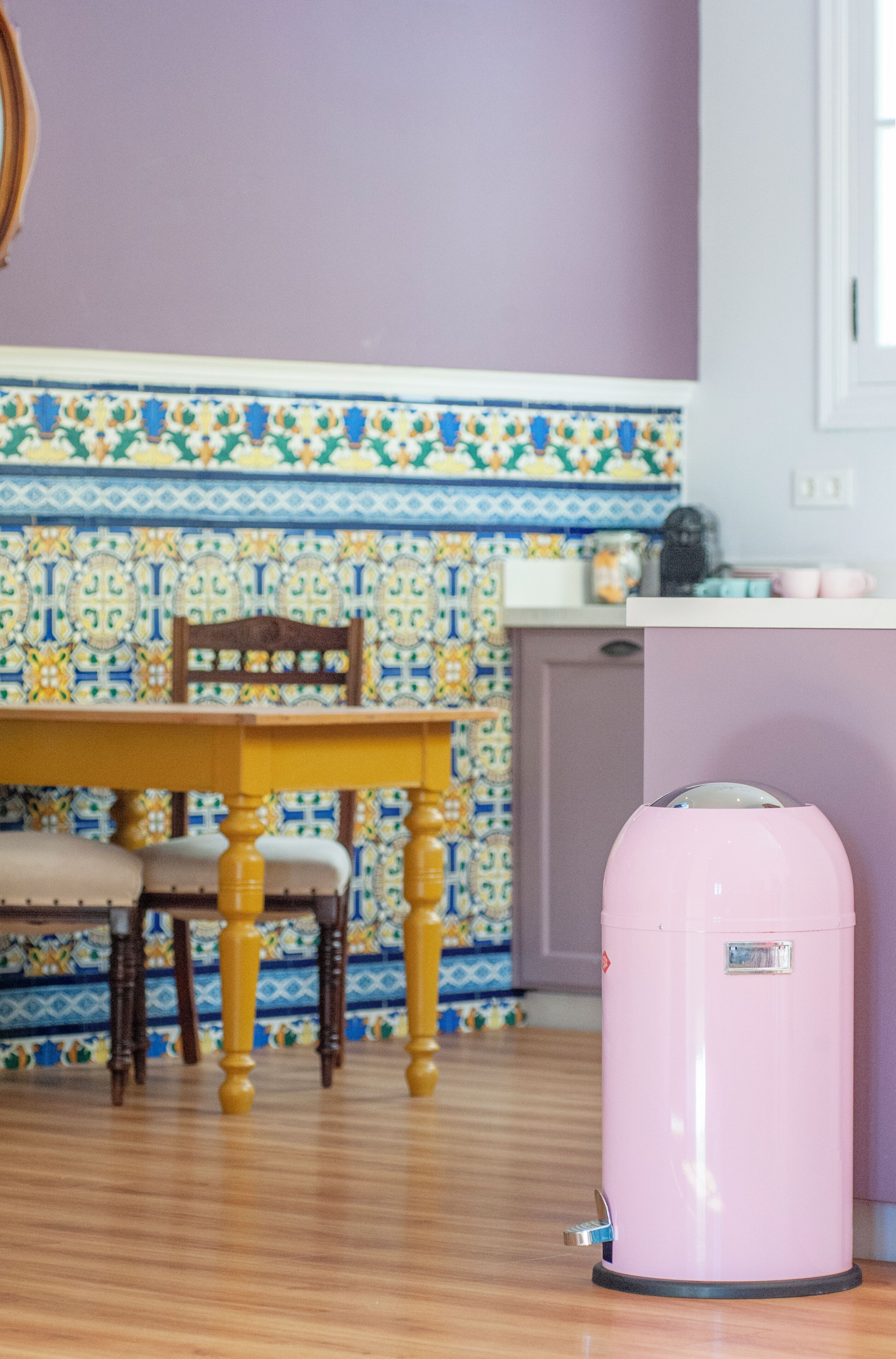 Une pièce avec un mur recouvert de carreaux décoratifs, une table en bois avec des chaises et une poubelle rose.