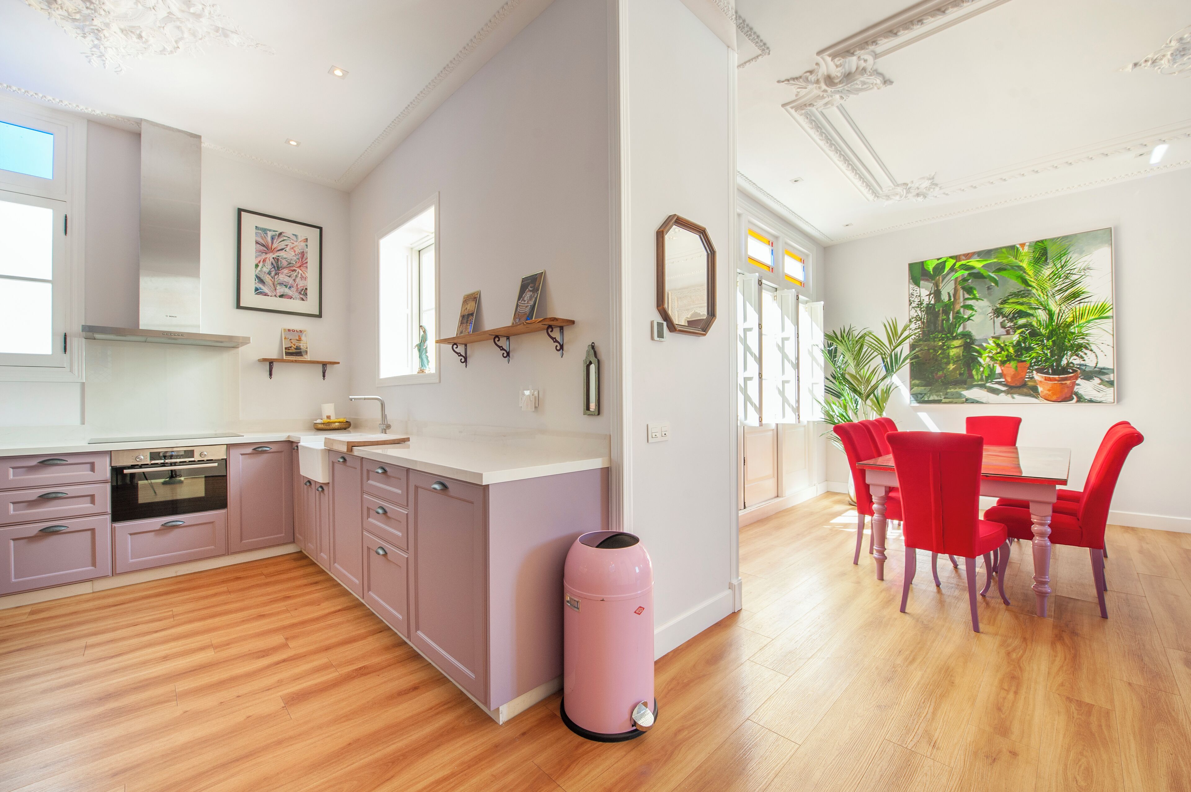 A modern kitchen with wooden flooring, a pink trash bin, and a dining area with red chairs and a large window.