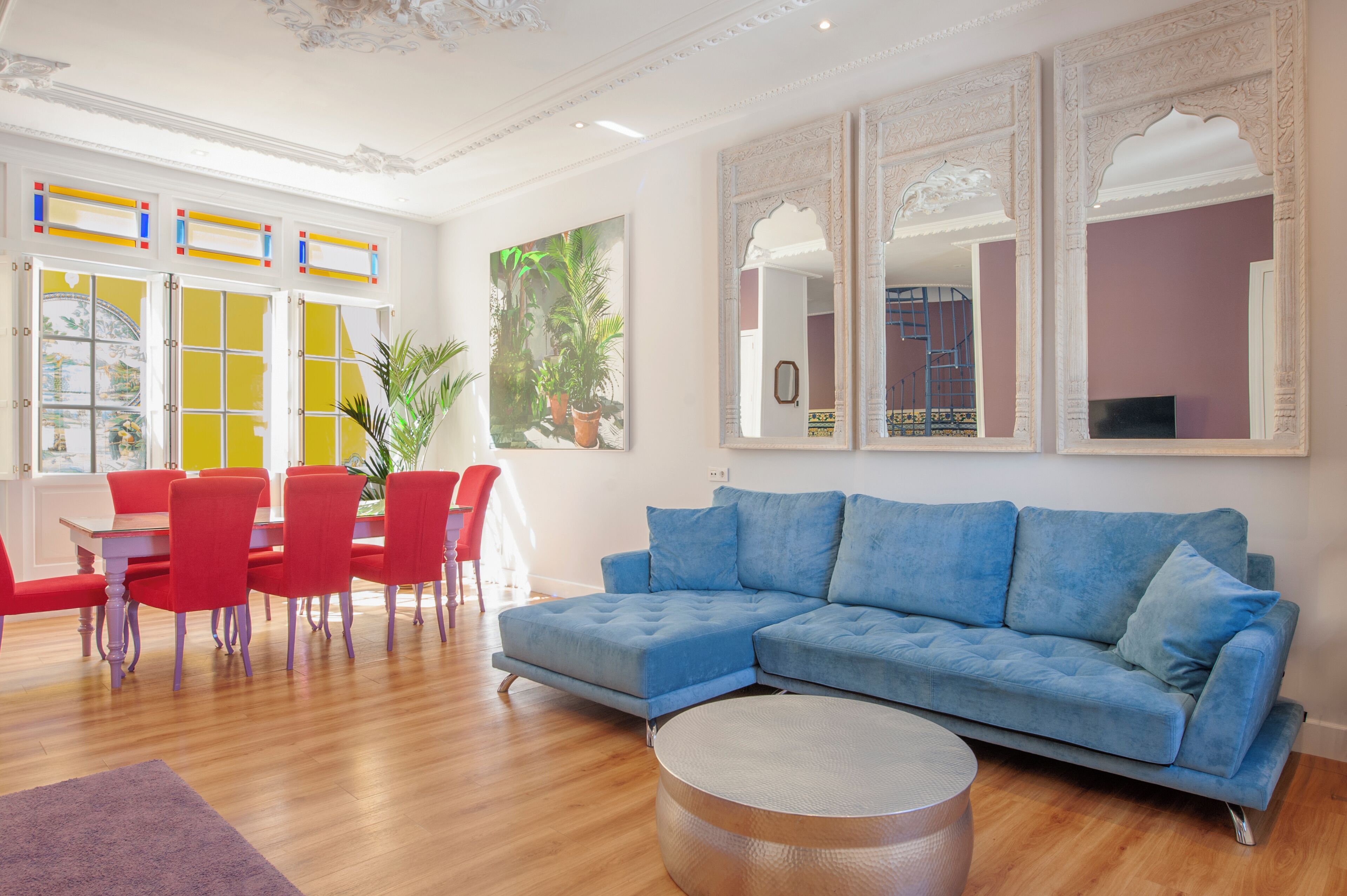 A modern living room with a blue sectional sofa, a dining table with red chairs, and a large mirror with ornate frames.
