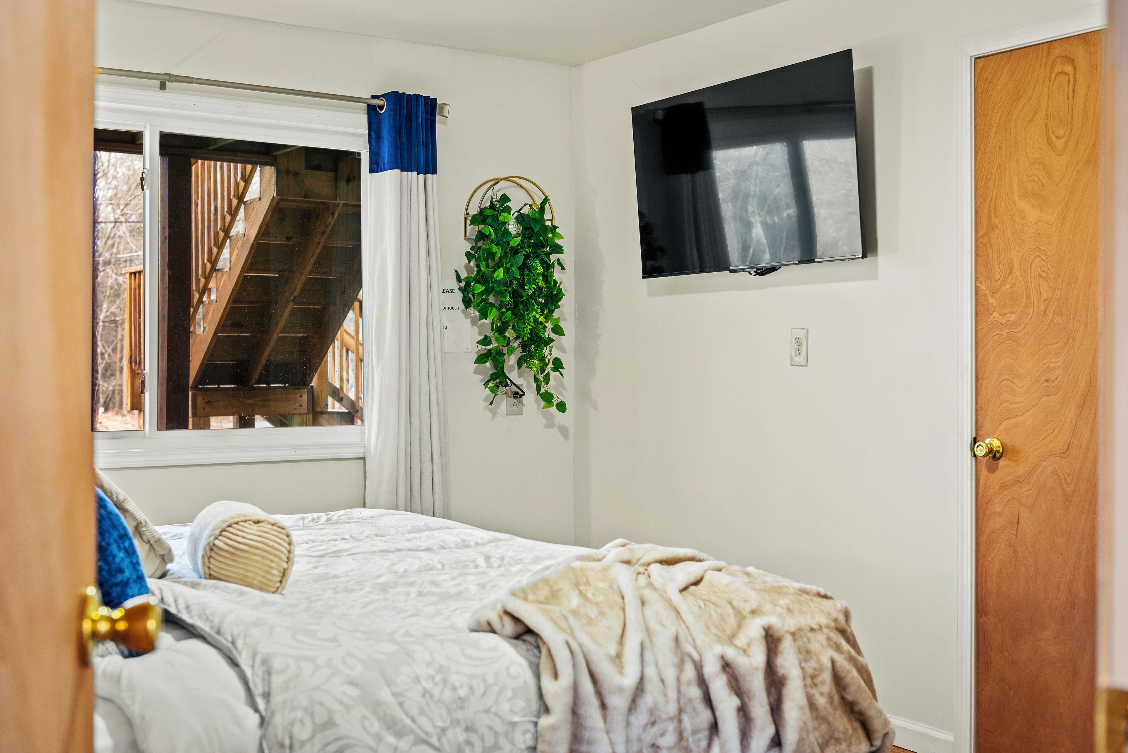 A bedroom with a bed, a TV mounted on the wall, a window with curtains, and a wooden door.