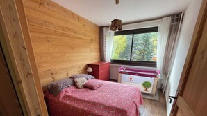 A bedroom with a wooden headboard, a bed with a floral bedspread, a nightstand with a lamp, and a window with a view of trees. - Chantemerle T2, 49m2, south-facing,   (Saint-Chaffrey)