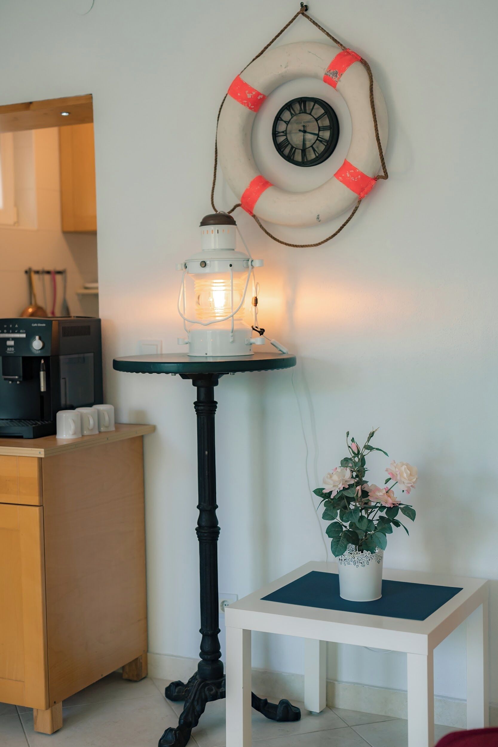 Una habitación pequeña con una cafetera, una mesa con lámpara, una planta en maceta y un reloj de pared.