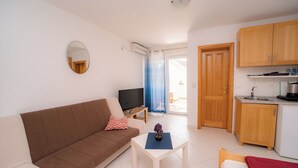 Ein modernes Wohnzimmer mit einem braunen Sofa, einem kleinen weißen Tisch und einem Fernseher. Im Hintergrund ist eine Küche mit Holzschränken und einem Spülbecken zu sehen.