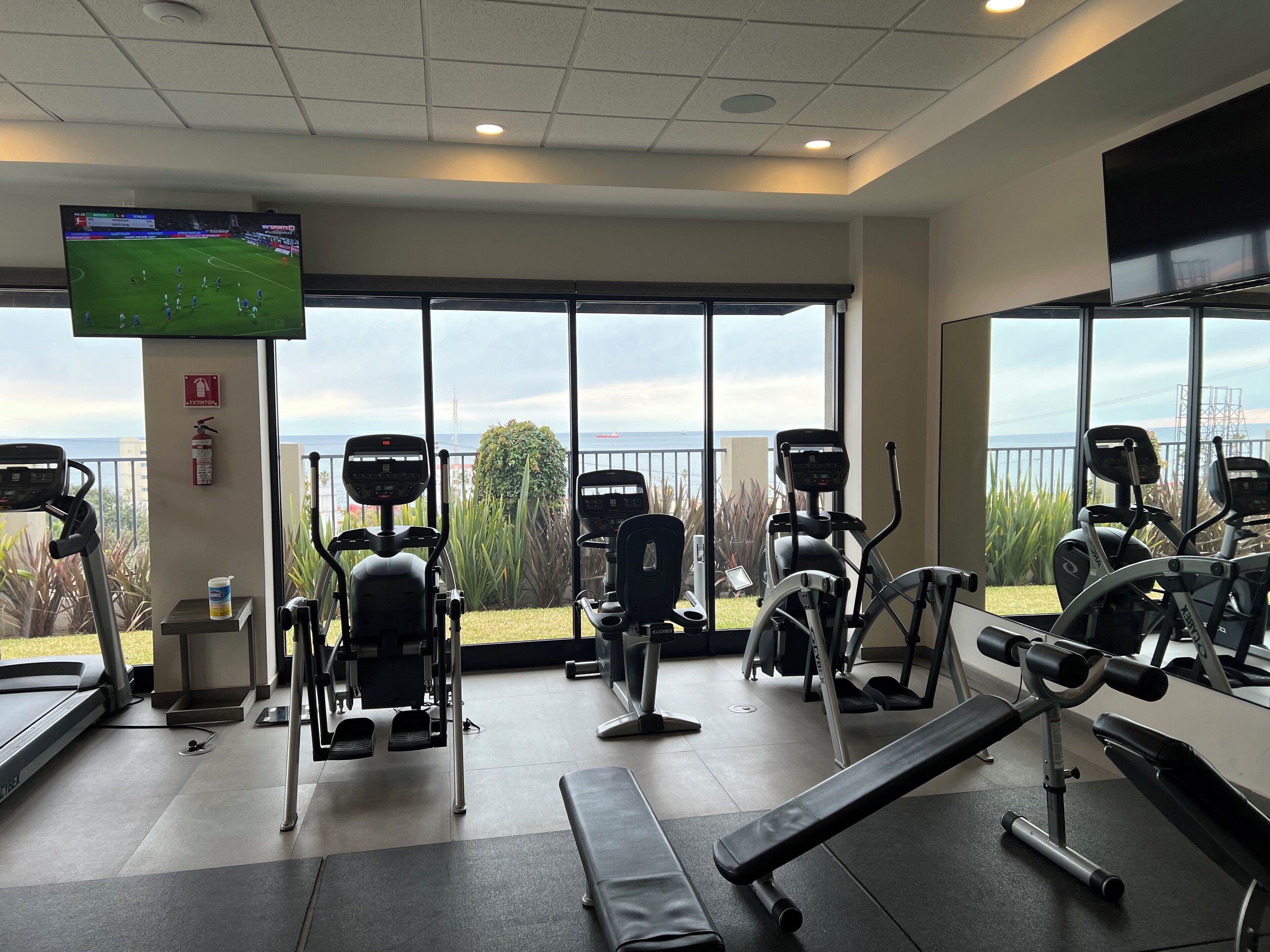 A gym with various exercise equipment, a television displaying a sports game, and large windows offering a view of the sea.