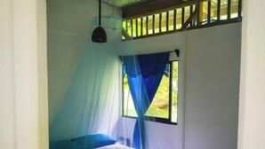 Uma cama de solteiro com mosquiteiro, soalho de madeira e janela com cortinas azuis.