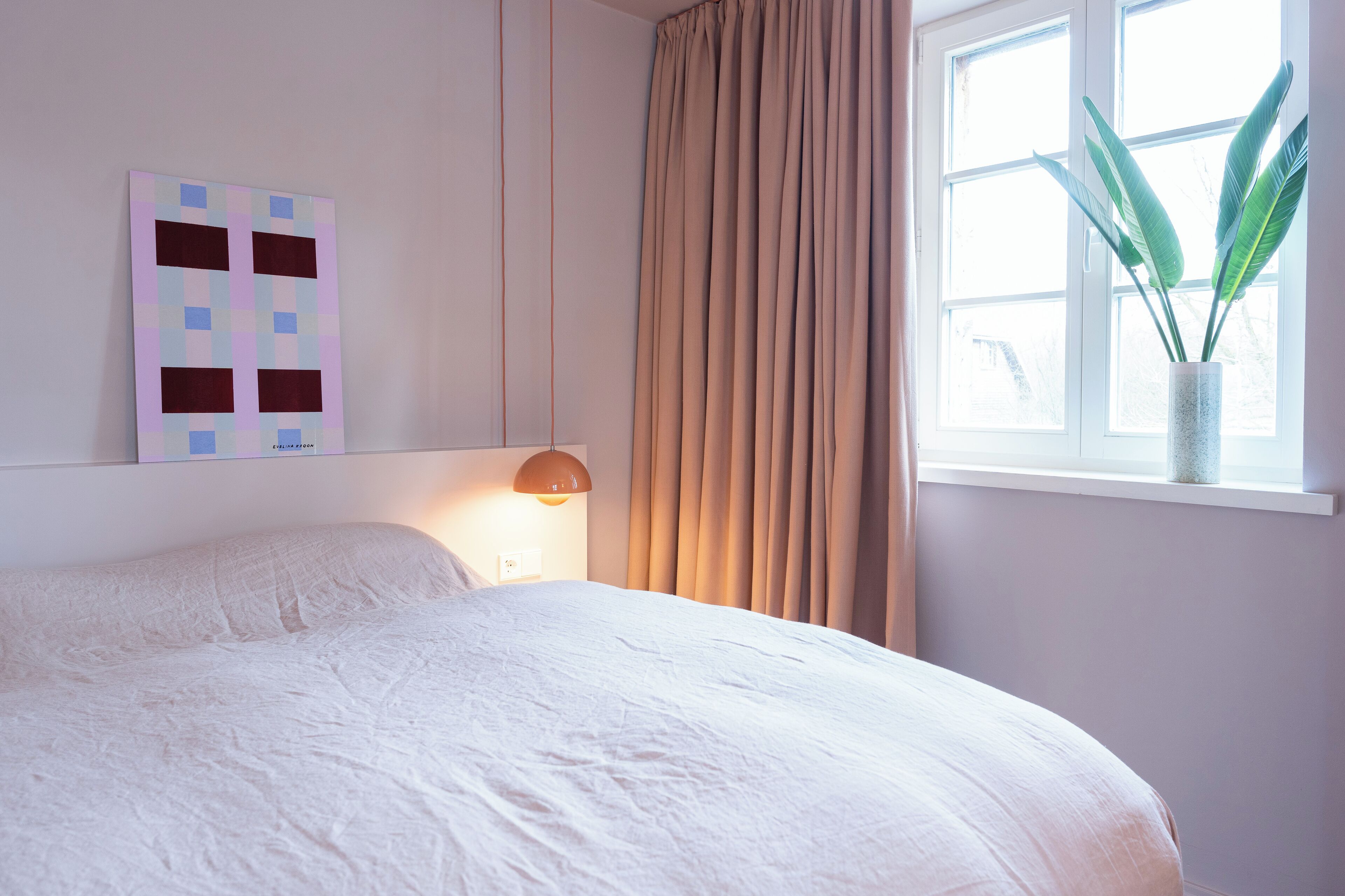 A neatly made bed in a room with a large window, a wall-mounted artwork, and a potted plant.