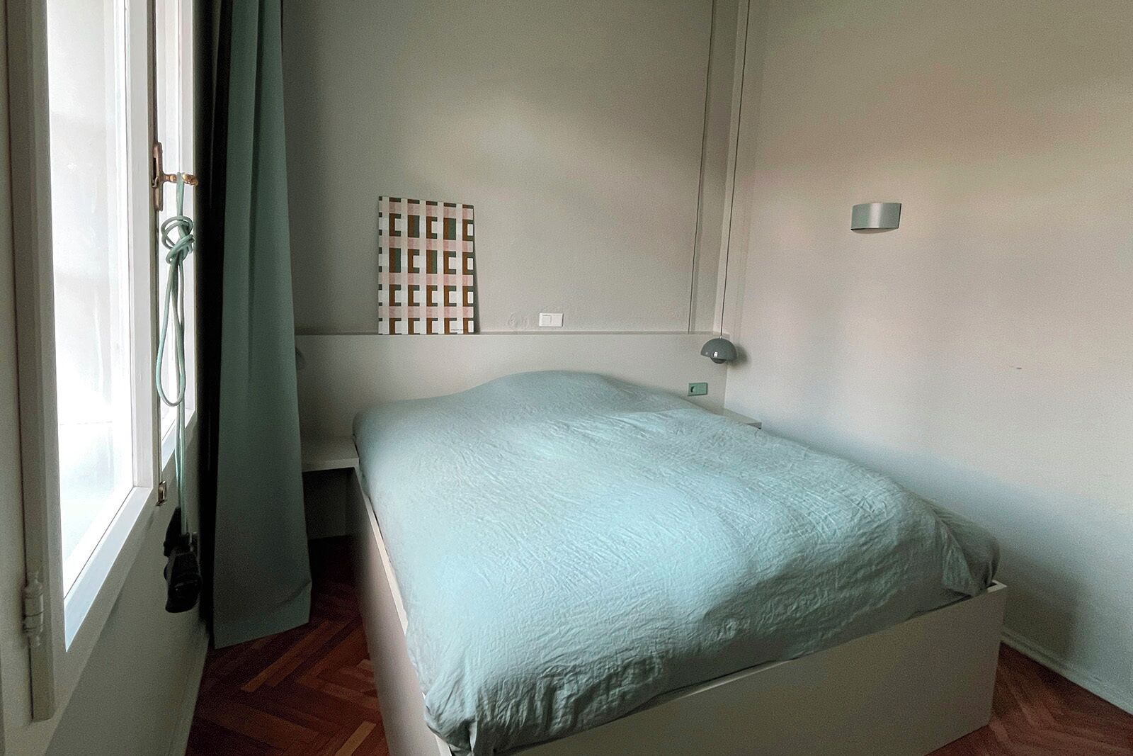 Una habitación pequeña con cama individual, una cama sencilla, una ventana con cortinas, una lámpara fijada a la pared y un estante con un objeto decorativo.