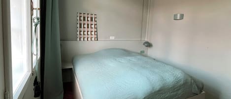 A small, single-bed room with a simple bed, a window with curtains, a wall-mounted light, and a small shelf with a patterned object.