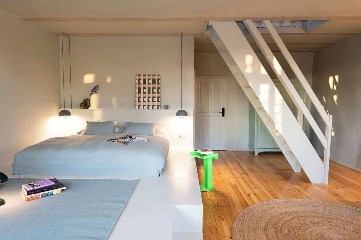 Ein Schlafzimmer mit einem Bett, Nachttischen, einer grünen Mülltonne, einer Treppe und einer Tür.