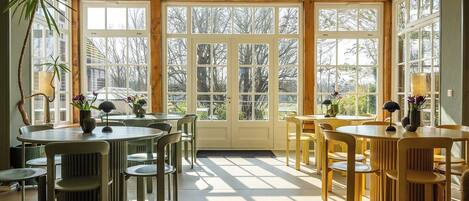 A bright, sunlit room with large windows, wooden beams, and multiple tables and chairs.
