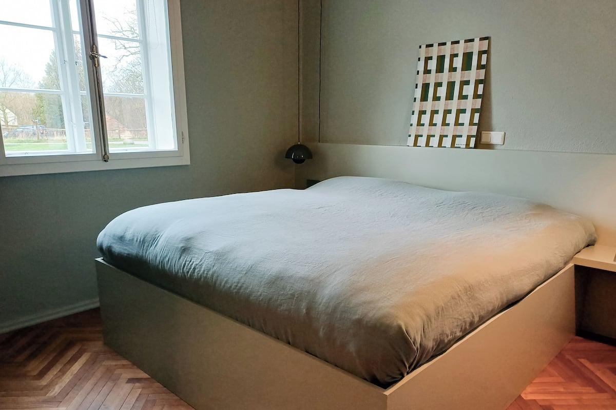 Ein Zimmer mit einem Bett, einem Fenster, einer Wandleuchte und einem Parkettboden mit Fischgrätenmuster.