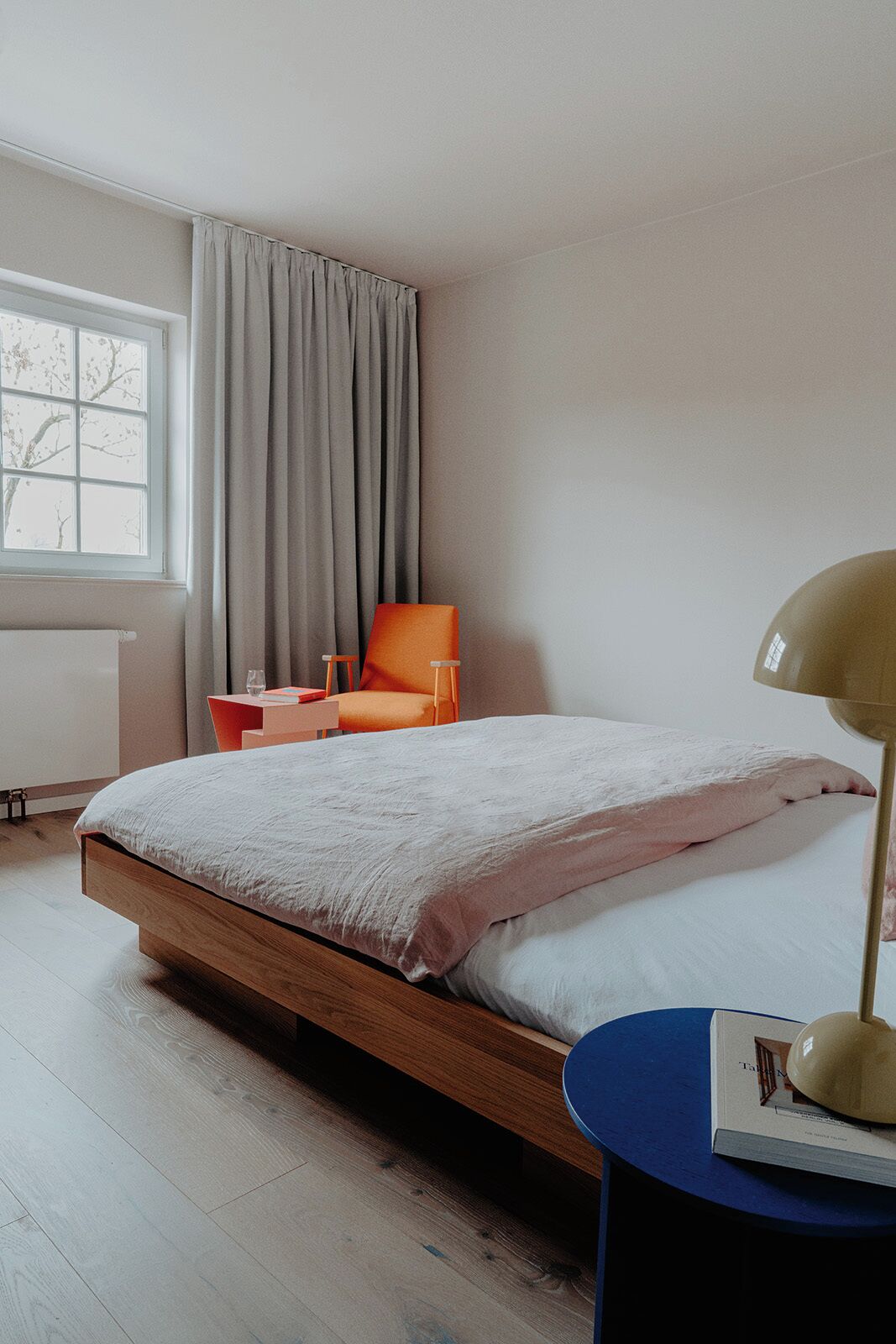 A bedroom with a wooden bed, a desk, an orange chair, a window with curtains, and a lamp on a side table.