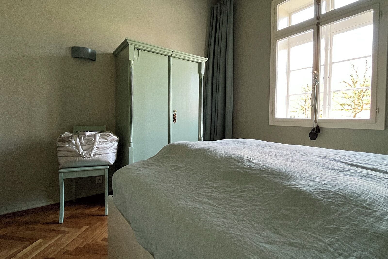 A bedroom with a bed, a wardrobe, a window with curtains, and a bag on a chair.