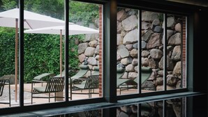 Een terras met buitenmeubilair, een stenen muur en uitzicht door grote ramen.