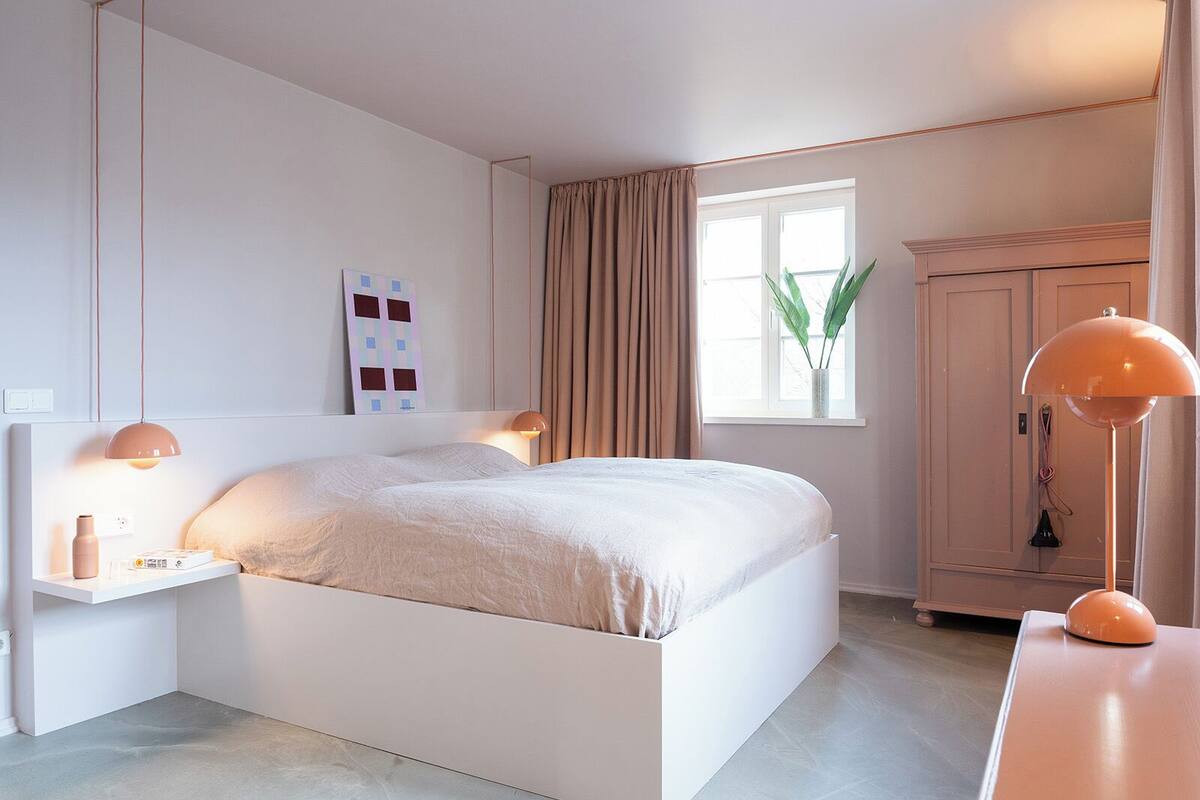 Ein schlicht eingerichtetes Schlafzimmer mit Bett, Nachttischlampen, einem Beistelltisch, einem Fenster mit Vorhängen und einer Pflanze.