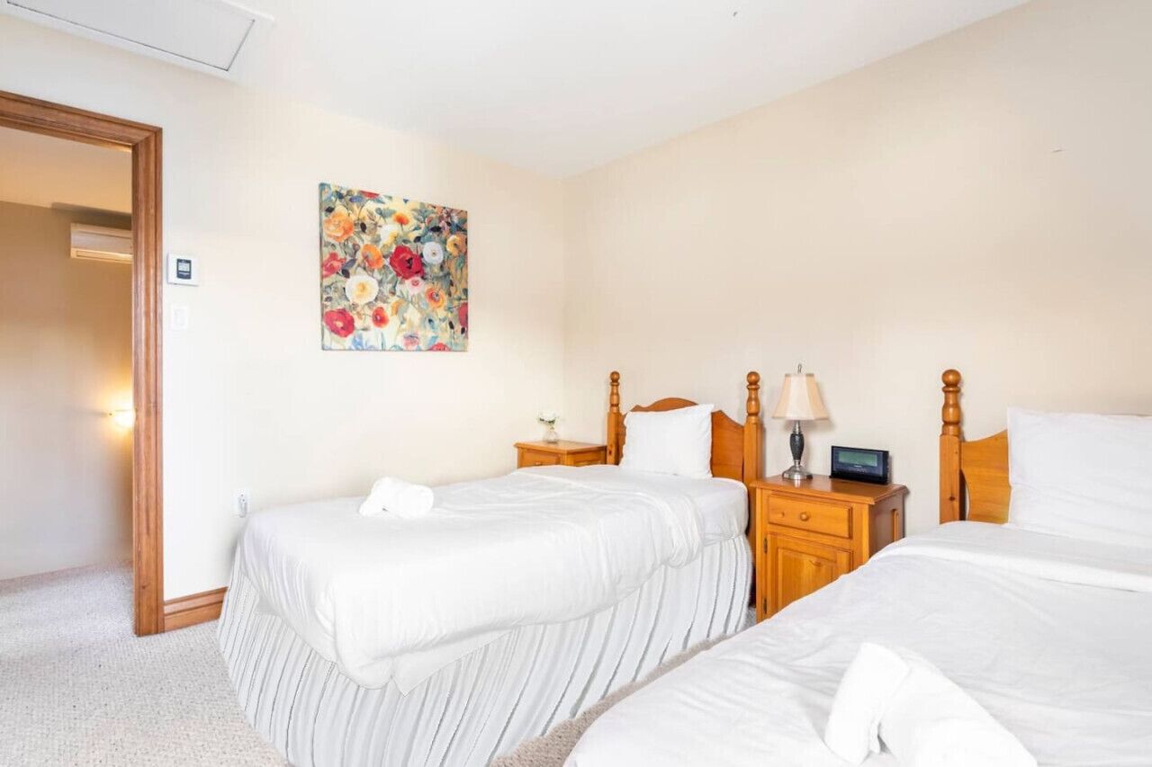 Habitación de hotel con dos camas, cabecera de madera, mesita de noche con lámpara y un cuadro con motivos florales en la pared.