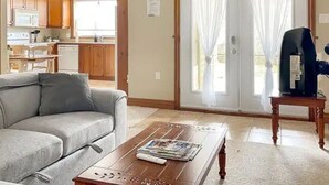 Ein Wohnzimmer mit grauem Sofa, einem Couchtisch mit Zeitschriften und einer Fernbedienung, einem Fernseher auf einem Holzboden und einer Glasschiebetür, die auf einen Balkon führt.