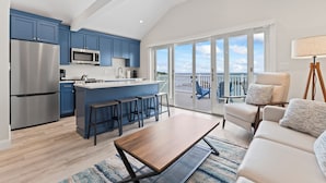 A modern kitchen with blue cabinets, a stainless steel refrigerator, and a microwave. There's a wooden dining table and a sofa with a lamp.