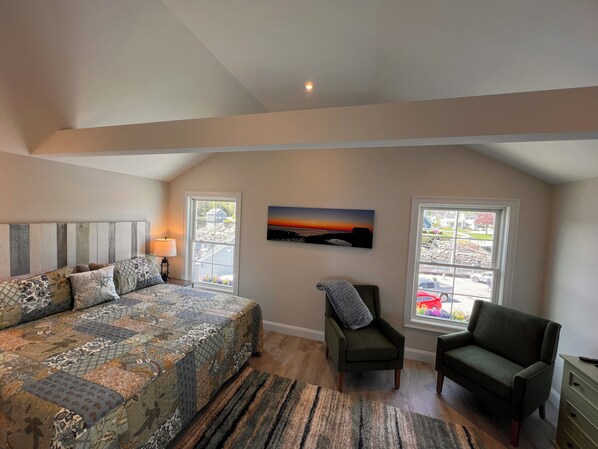 Un dormitorio con una cama grande, dos ventanas, una silla y un cuadro en la pared.