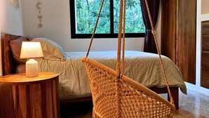 A bedroom with a bed, a hanging chair, a bedside table with a lamp, and a window with a view of trees.