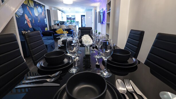 Mesa de comedor dispuesta con platos negros, cubiertos y copas de vino, rodeada de sillas tapizadas en negro.