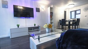 Una sala moderna con un televisor de pantalla plana montado en la pared, una mesa de centro de vidrio, un mueble blanco para entretenimiento y un comedor con mesa y sillas.