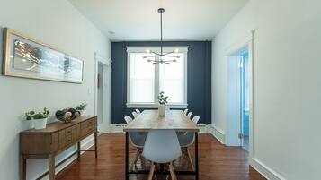 A dining area with a wooden table, white chairs, a pendant light, a wall-mounted painting, and a potted plant.