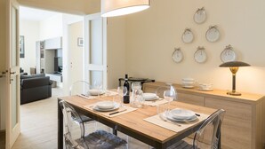 Un comedor con una mesa de madera dispuesta para cuatro personas, sillas traslúcidas, una botella de vino y una cafetera. Se observa un colgador de platos con platos.