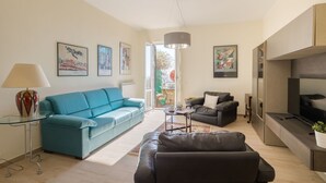 Un salón con un sofá azul, un sillón de cuero negro, una mesa de centro de vidrio y un televisor. Hay cuadros enmarcados en la pared y una lámpara sobre una mesita auxiliar.