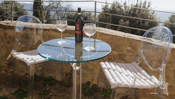 Una mesa de vidrio con dos sillas de plástico transparente, una botella de vino y dos copas de vino en una terraza con vista al mar y árboles.