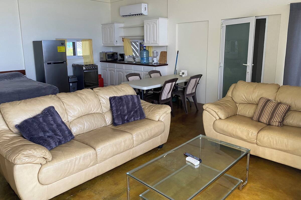 Een woonkamer met twee beige leren banken, een glazen salontafel, en een keuken met witte kasten en apparatuur.
