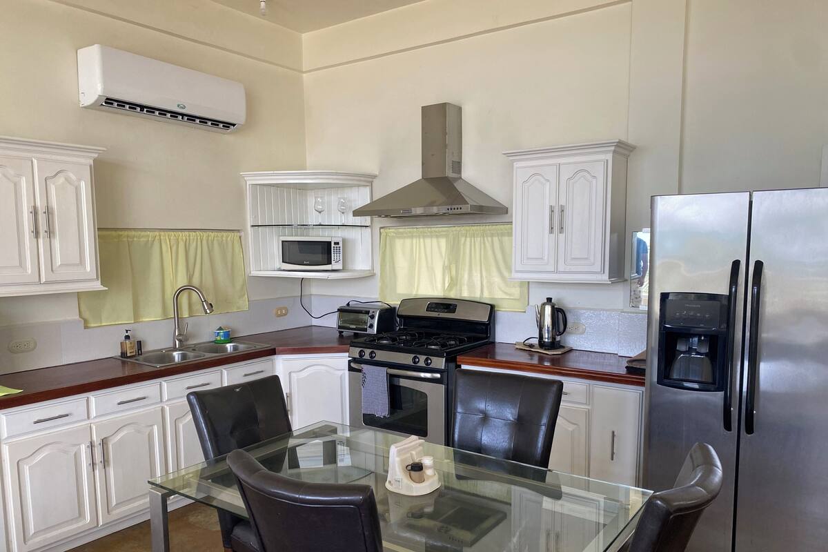 Een moderne keuken met witte kasten, een roestvrijstalen koelkast, een magnetron, een fornuis en een glazen eettafel met stoelen.
