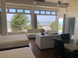 Habitación de hotel con cama, sofá, escritorio y televisor. Amplias ventanas con vista a la playa y palmeras.