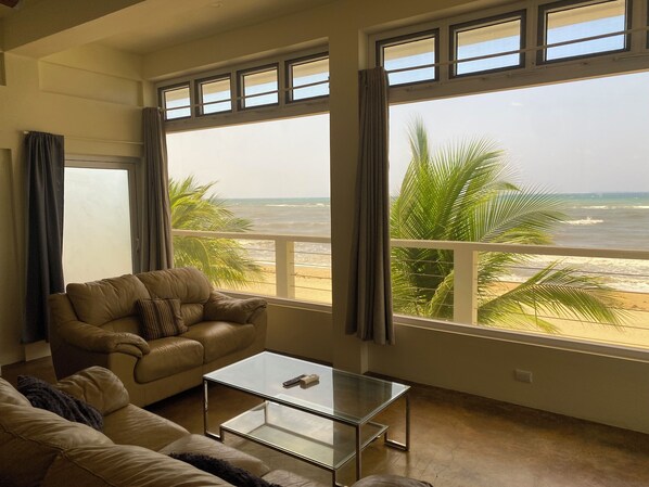 A living room with a sofa, a glass coffee table, and a view of the beach through large windows. - Trujillo Airport Beach Suites (Trujillo)
