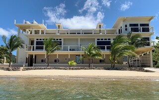 Una propiedad frente al mar de varios pisos con balcones, palmeras y una playa arenosa al frente.
