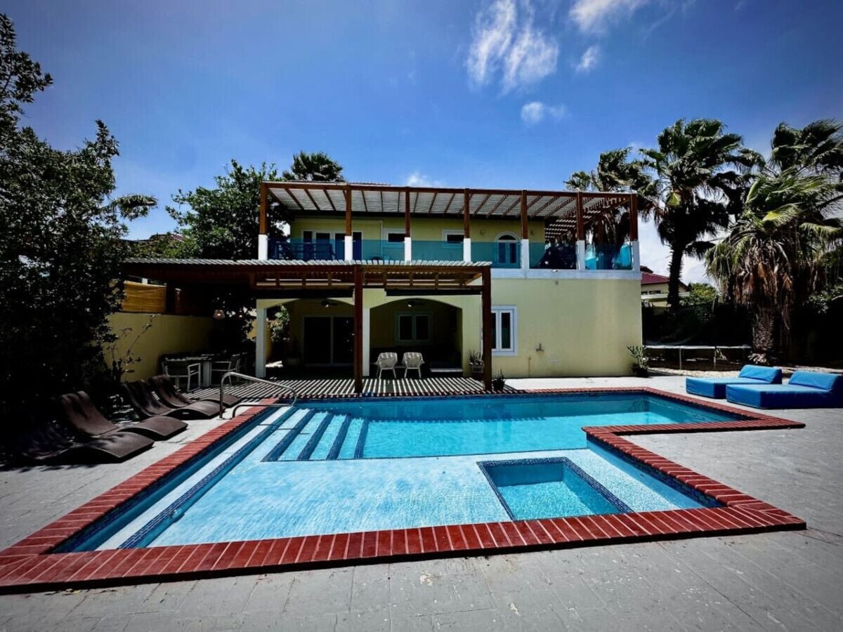 A swimming pool with a pergola and lounge chairs, surrounded by palm trees and a building with a balcony.