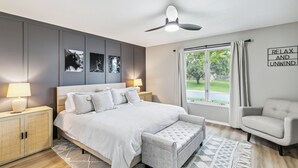 Een moderne slaapkamer met een bed, nachtkastjes, een bankje, een stoel, een plafondventilator en een raam met gordijnen.