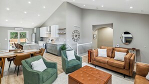 Een ruime woonkamer met een eettafel, groene fauteuils, een bruine bank en een keuken op de achtergrond.