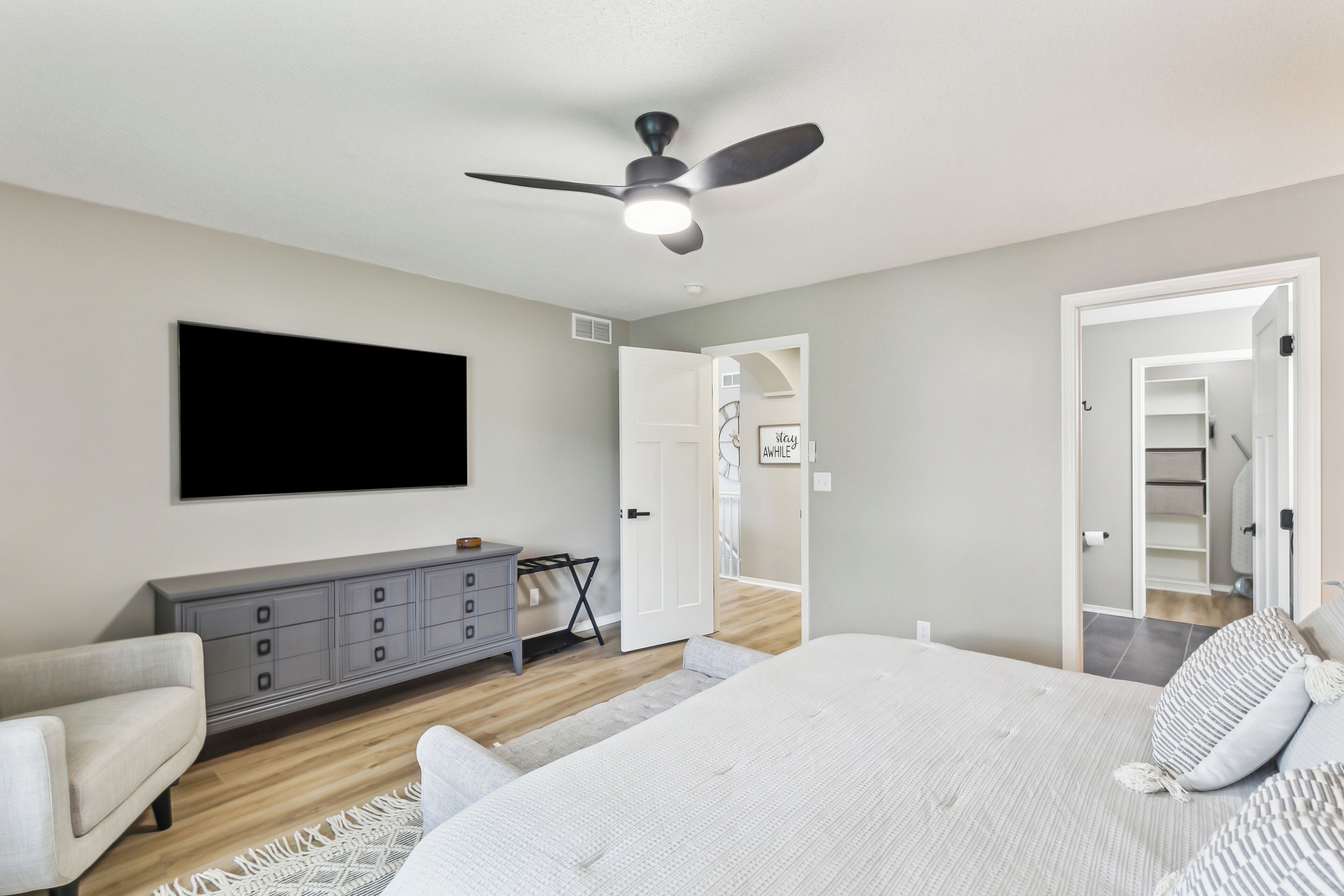 Quarto com cama, penteadeira, ventoinha no teto e TV fixada na parede.