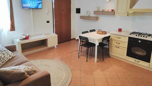 A compact living space with a sofa, a dining table with chairs, a kitchen area with a stove and oven, and a television mounted on the wall.
