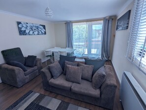 A living room with a sofa set, a dining table, and chairs. There is a large window with curtains, a chandelier, and a painting on the wall. - A&A Apartment Basildon with free parking  (Basildon)