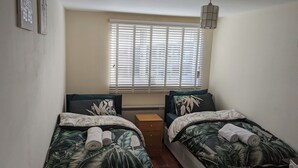 A small, well-lit bedroom with two single beds, a window with blinds, a bedside table, and a hanging light fixture. - A&A Apartment Basildon with free parking  (Basildon)